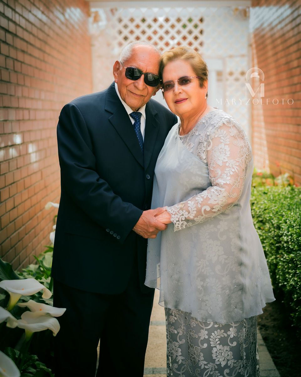 Pareja de adultos mayores de pie en un pasillo, están recargando sus cabezas y tomándose de las manos.