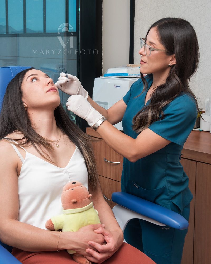 Doctora en sala de exploración aplicando gotas oftalmológicas en paciente sentada. La paciente tiene sobre sus manos un peluche.