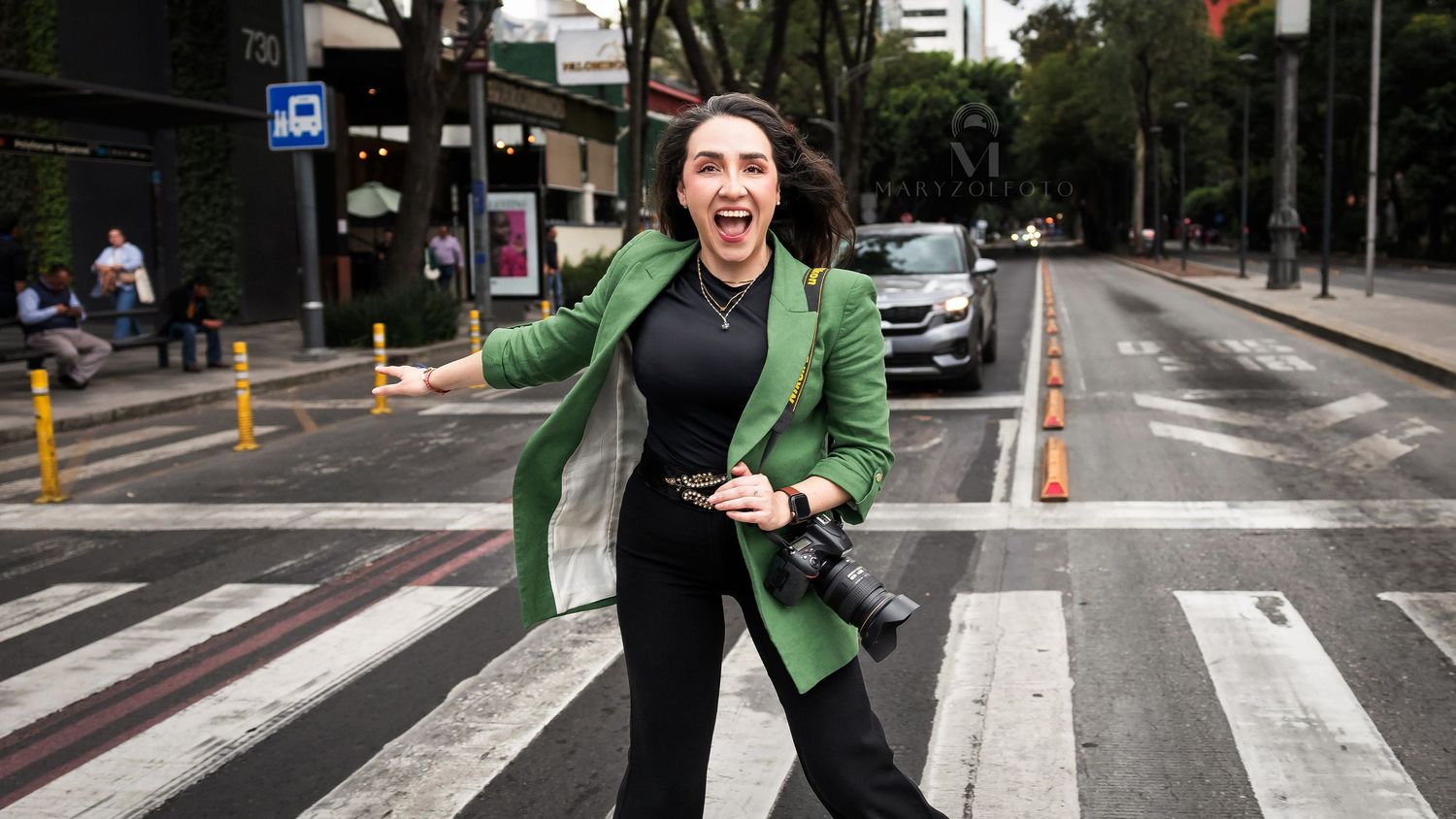 Fotógrafa sonriendo cruzando un paso peatonal, cuelga sobre uno de sus hombros su cámara.