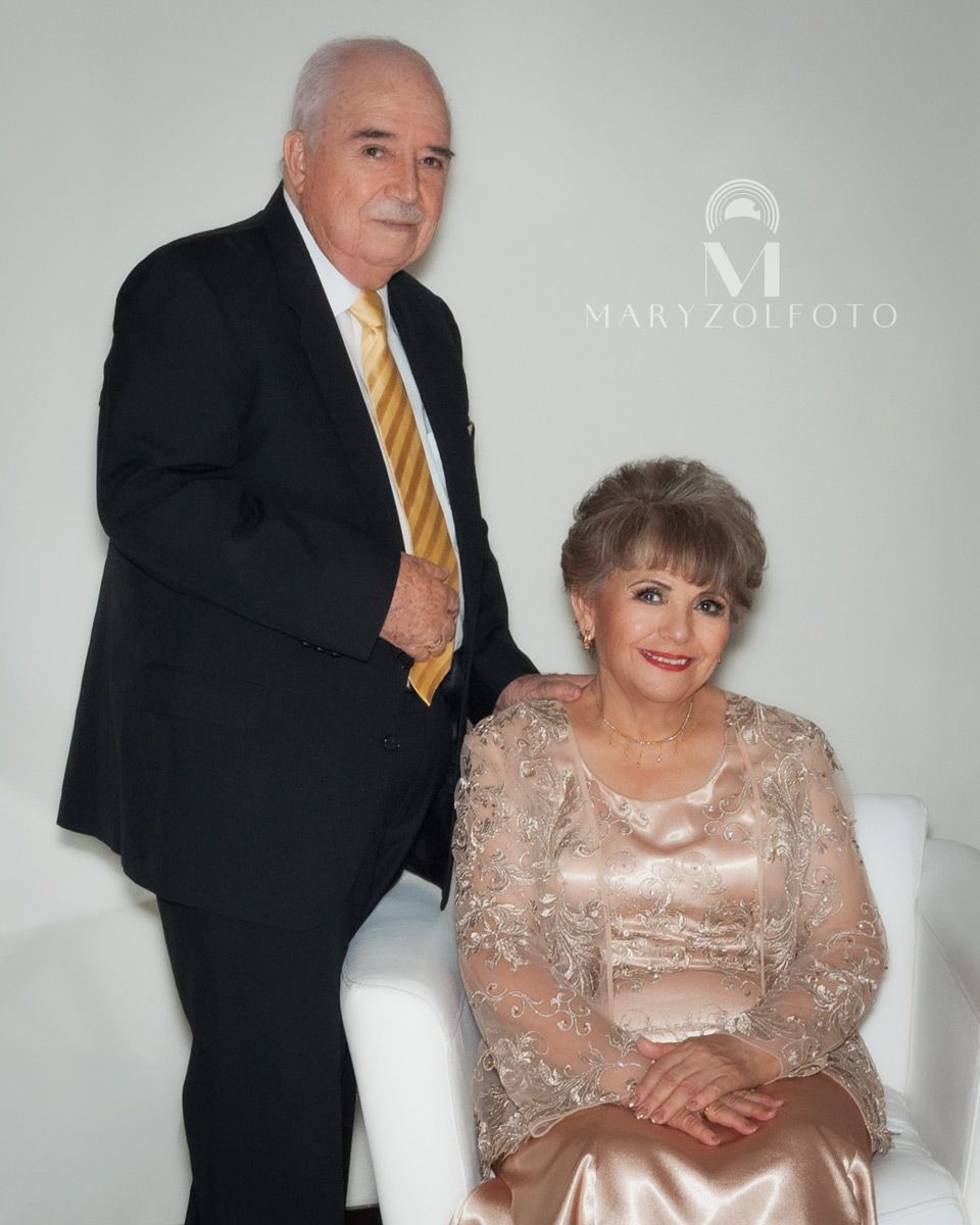 Pareja de adultos mayores sonriendo, vestidos formalmente. La mujer está sentada y el hombre de pie detrás de ella.