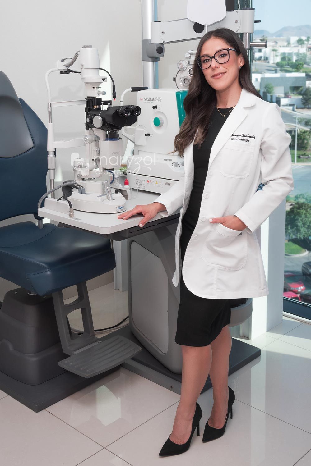Doctora sonriendo de pie en sala de exploración oftalmológica, usando vestido midi negro, tacones negros y bata médica.