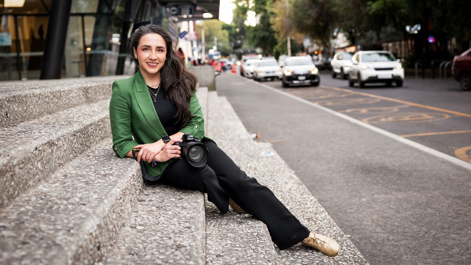 Fotógrafa feliz sentada sobre escaleras, viste de negro con un saco verde. Sobre su pierna descansa su cámara.