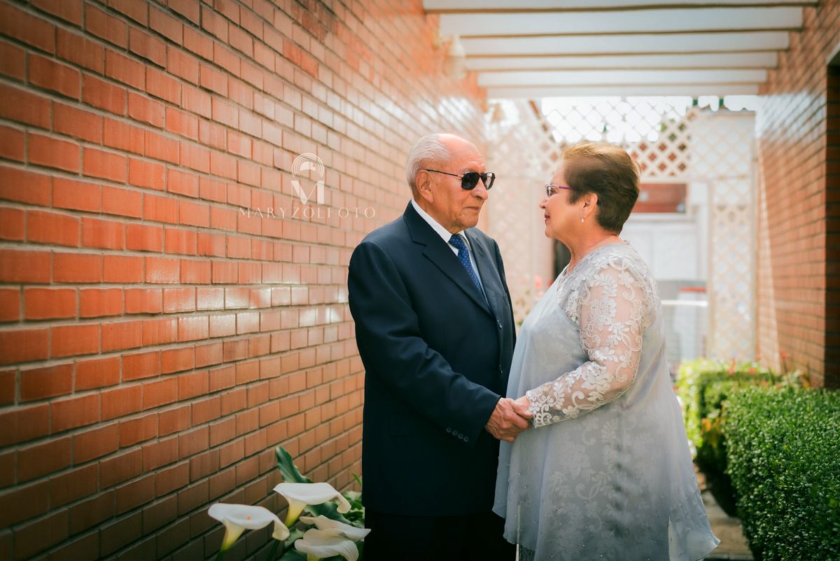 Pareja de adultos mayores de pie en un pasillo, se están uno frente al otro y están mirándose y tomándose de las manos.