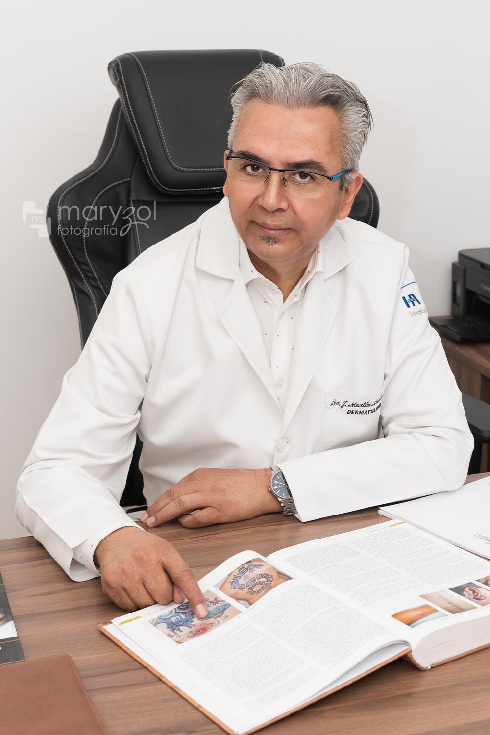 Doctor sonriendo sentando en su consultorio, tiene un libro abierto y con un dedo está señalando una parte del libro.