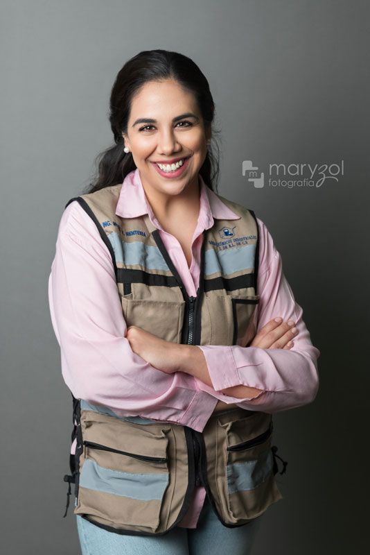 Retrato de estudio mujer profesionista sonriendo, usando blusa rosa y chaleco de seguridad café con franjas reflejantes.