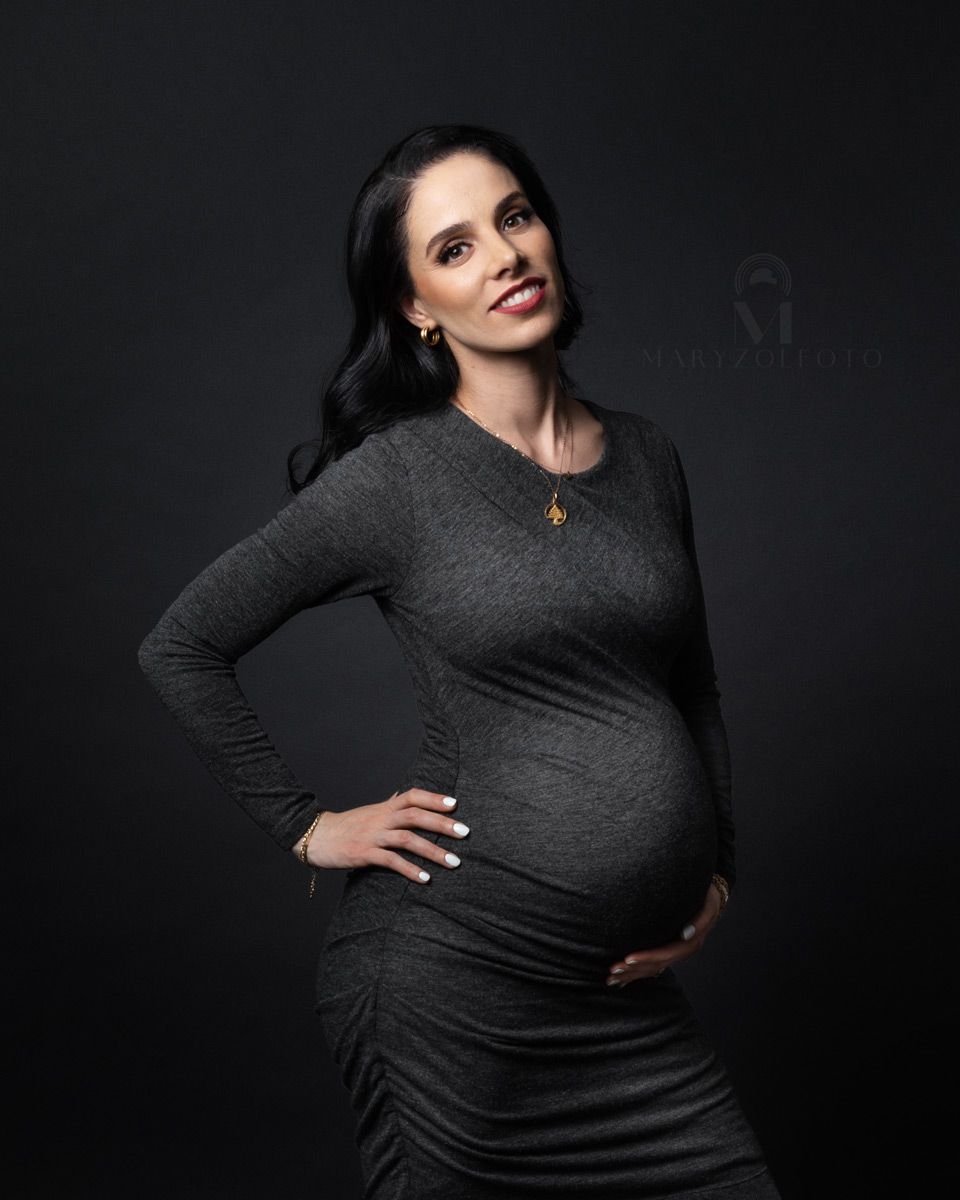 Retrato de estudio de mujer sonriendo embarazada, usa un vestido gris oscuro y esta sosteniendo su vientre con una mano.