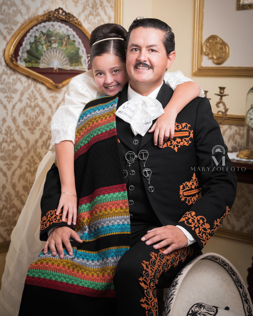 Hombre sentado usando traje tradicional de charro, sobre él se recarga una niña con vestido blanca que lo abraza por detrás.