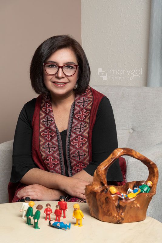 Mujer profesionista sentada en sillón, frente a ella hay una mesa con figuras Playmobil.