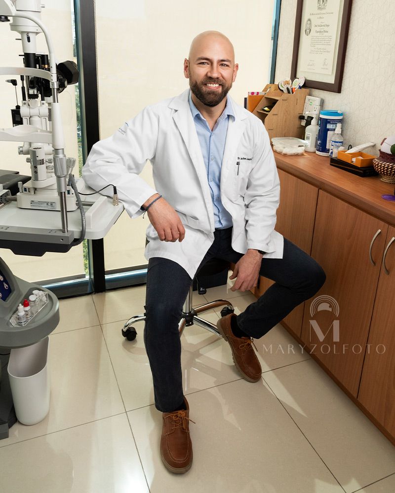 Doctor sentado en sala de exploración oftalmológica, viste casual con bata médica.