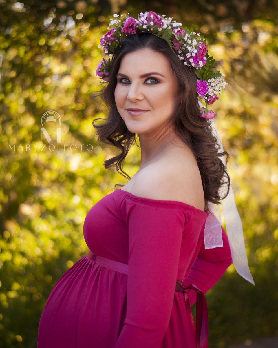 Mujer embarazada sonriendo, cuerpo está de lado y su rostro mira hacia la cámara. Usa vestido rosa y una corona de flores.