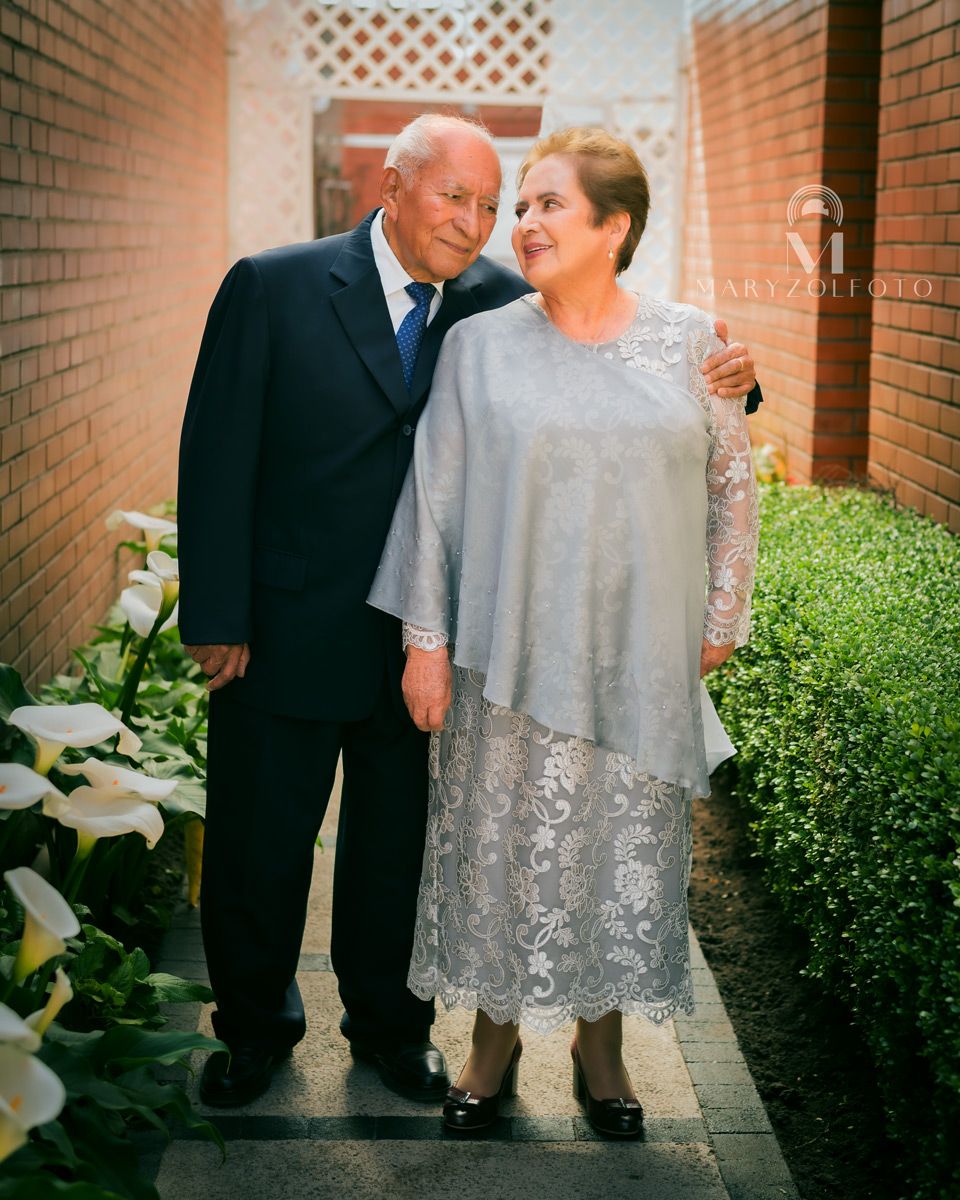 Pareja de adultos mayores de pie en un pasillo, están mirándose de lado y sonriendo. El hombre pasa su brazo por detrás de la mujer.