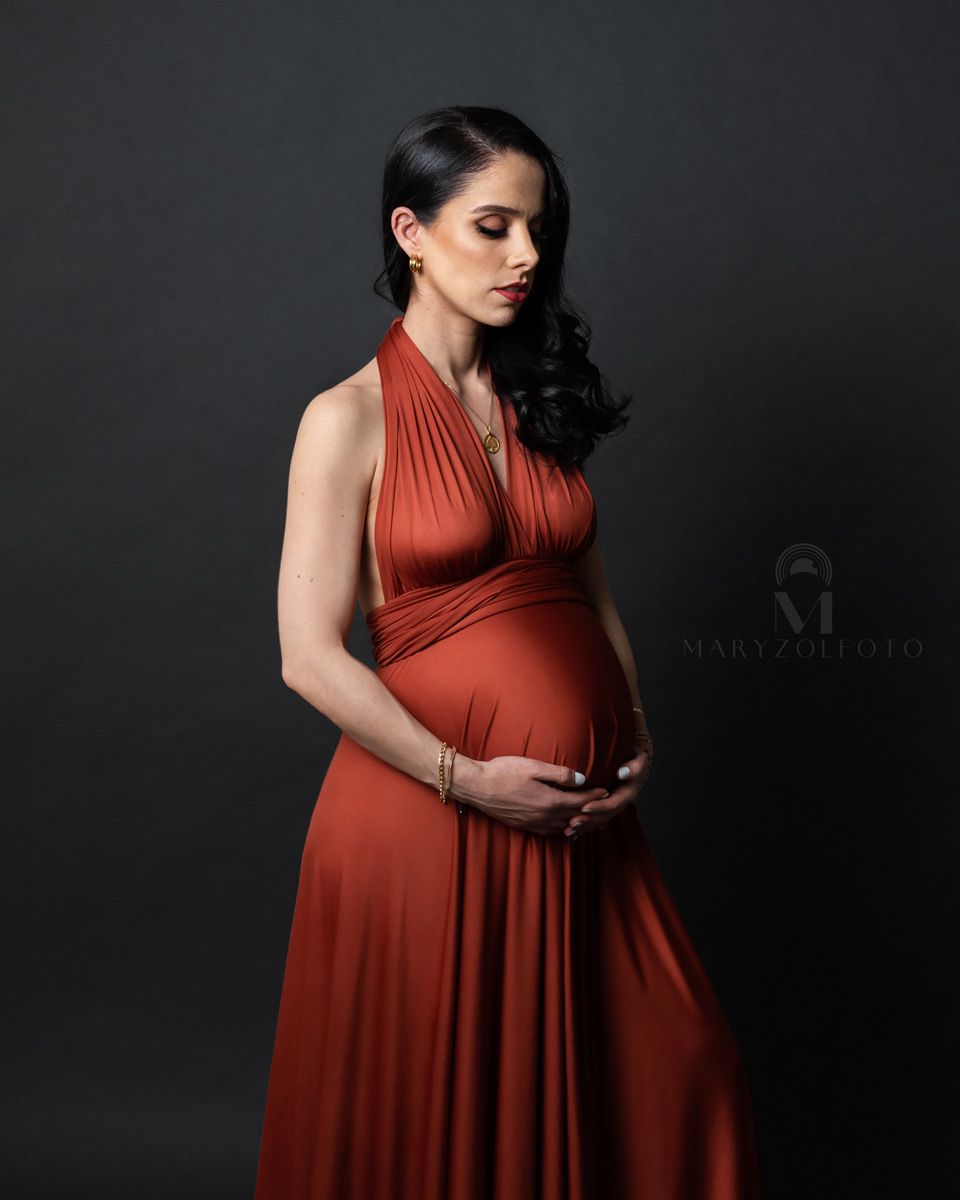 Retrato de estudio de mujer embarazada, usa un vestido naranja y esta mirando su vientre y lo sostiene con ambas manos.