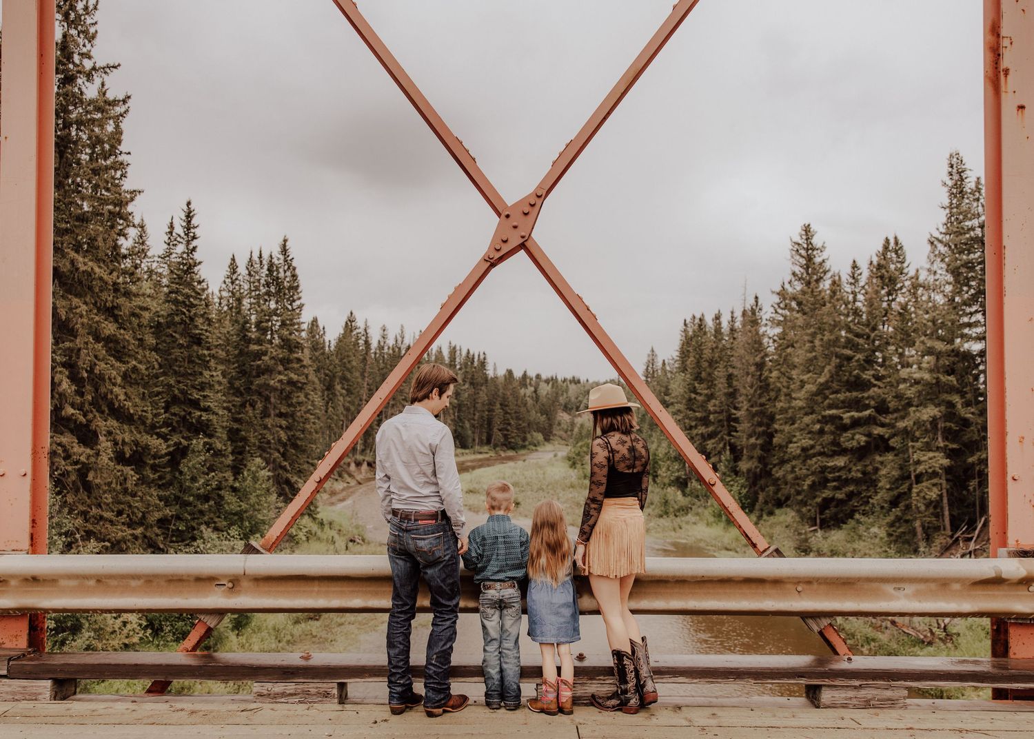 Family of four standing on a rustic red bridge overlooking a lush pine forest in the mountains.