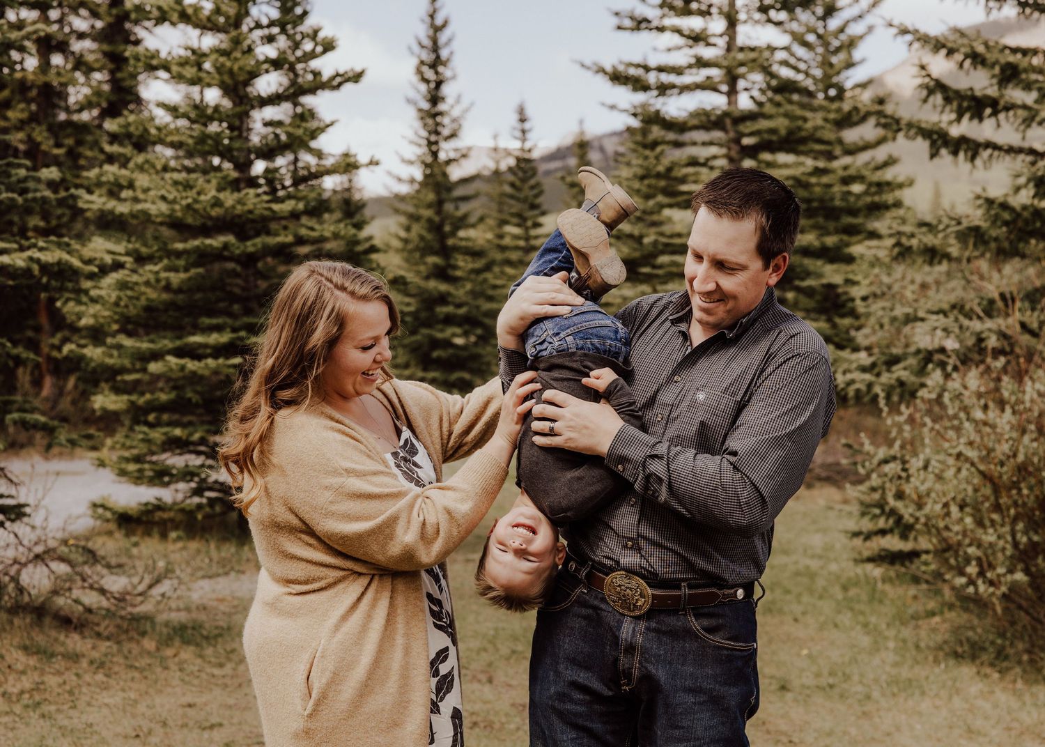 A couple stands outdoors among pine trees, holding their baby together in a warm family portrait.