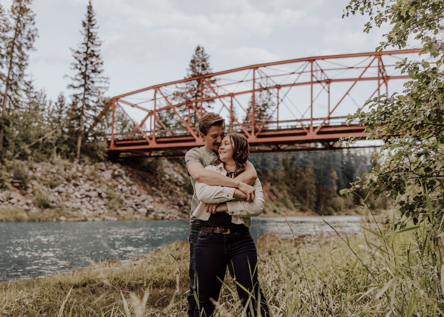 Couple enjoys a romantic moment near a red metal bridge in a scenic outdoor setting during golden hour.