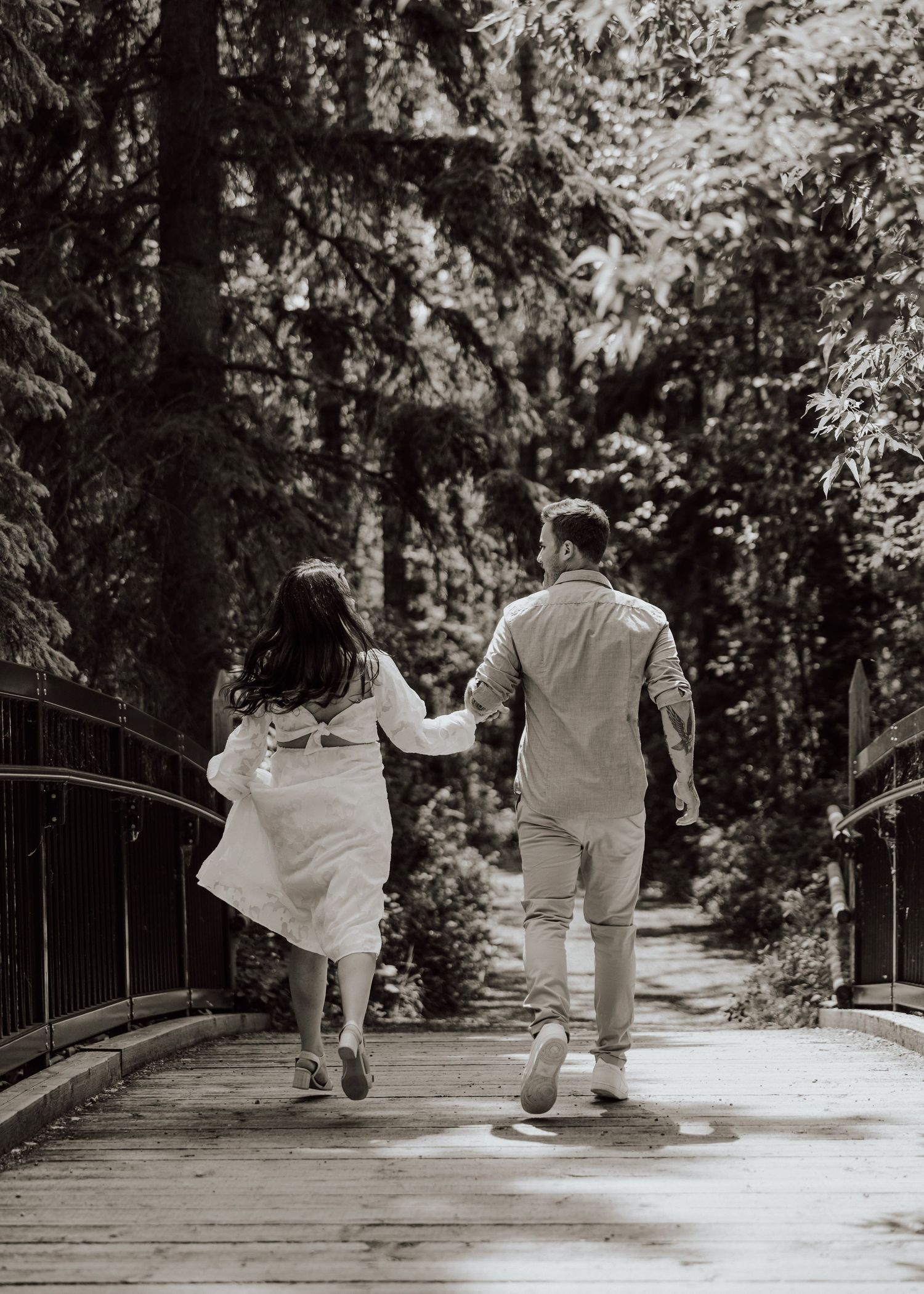 A couple in white outfits holds hands while joyfully running across a wooden bridge in a lush forest setting.