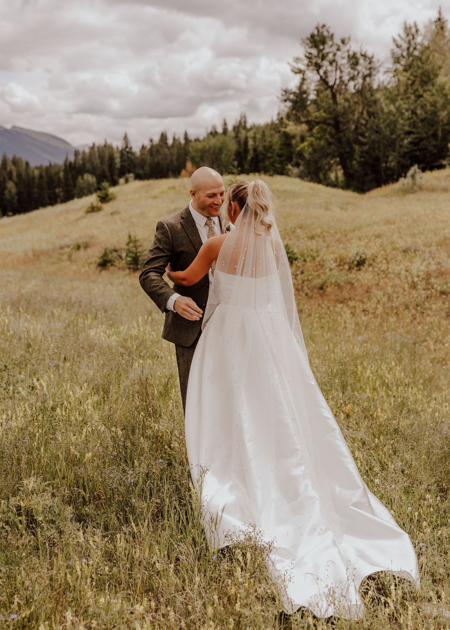A couple shares an intimate moment on a mountainside meadow with evergreen trees and cloudy skies in the background.