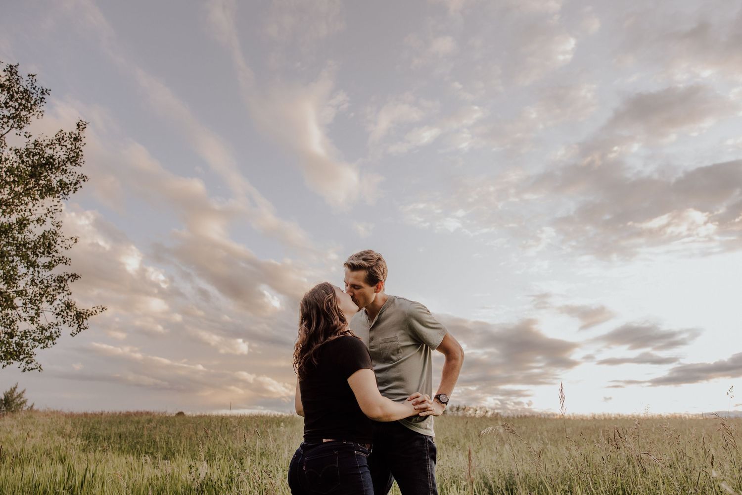 A couple embraces in a golden field during sunset while clouds paint the sky in warm hues.