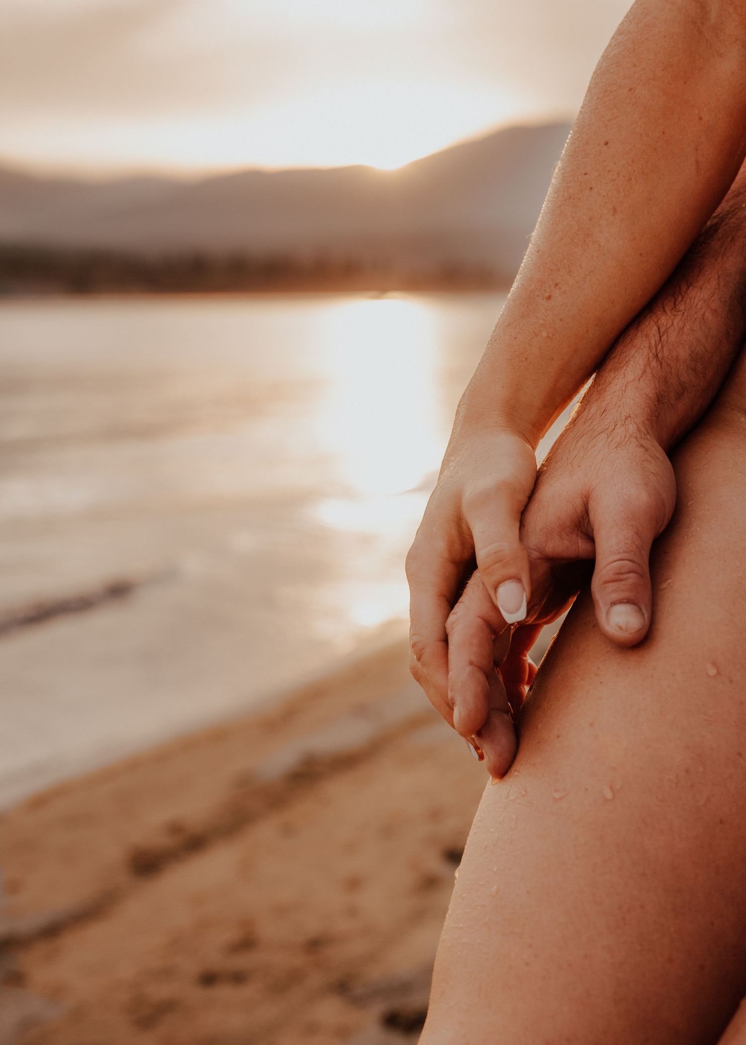 Close-up of two people holding hands on a sandy beach at golden sunset with mountains in background.