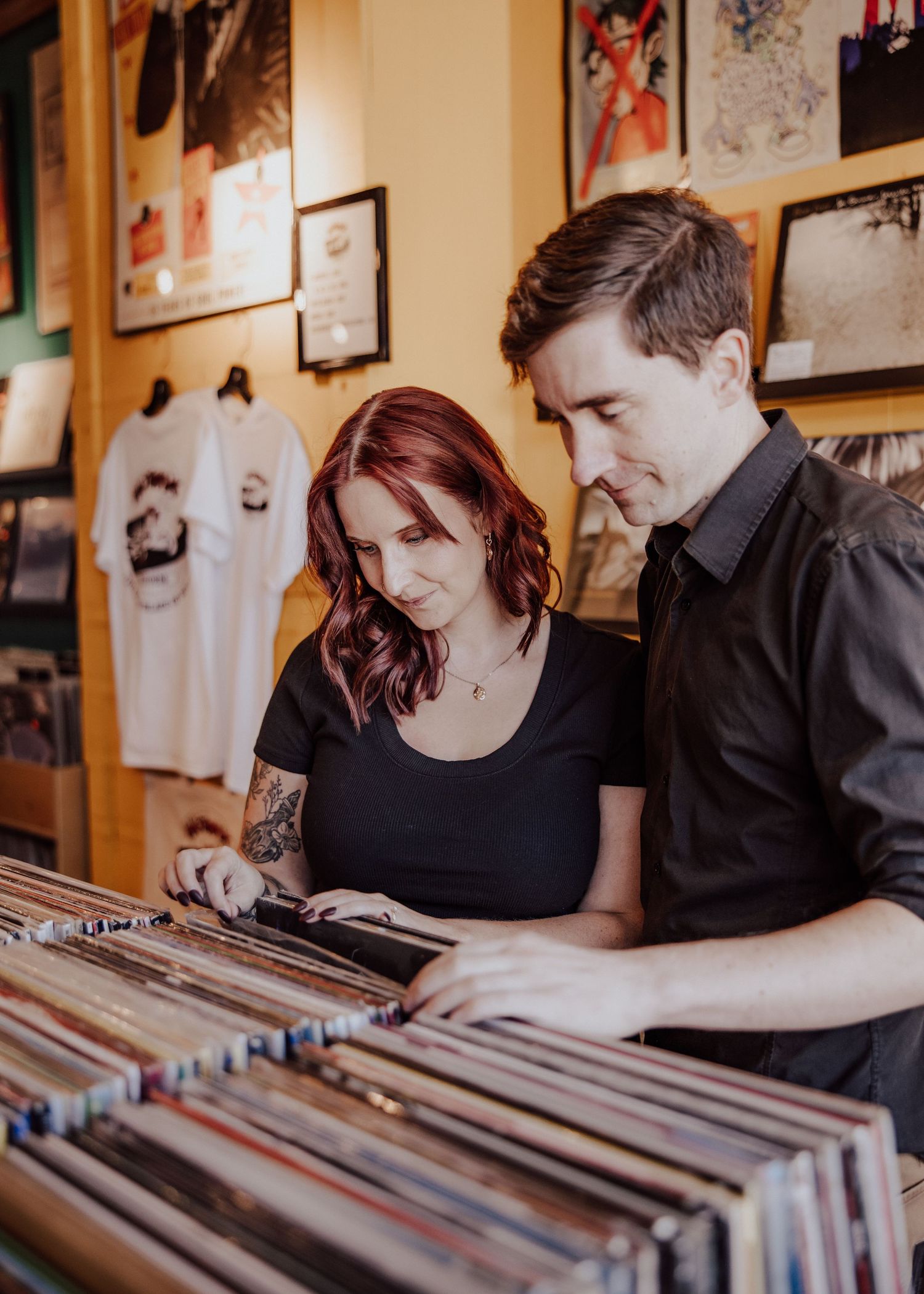 A couple browses vinyl records together in a cozy music shop, the woman with red hair and tattoos leaning in curiously.