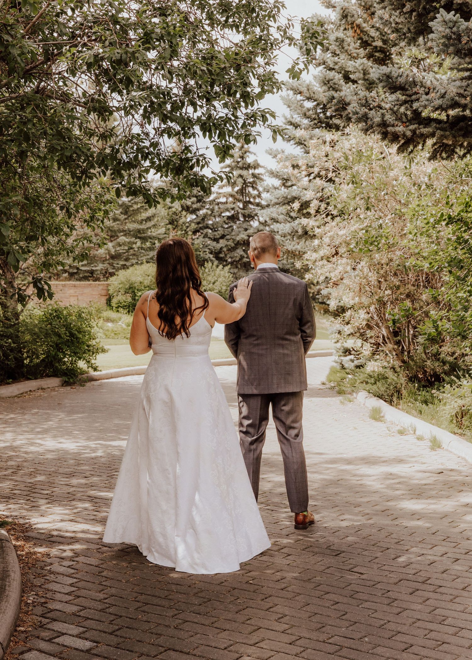 A couple in wedding attire walking together on a brick pathway through a lush garden setting.