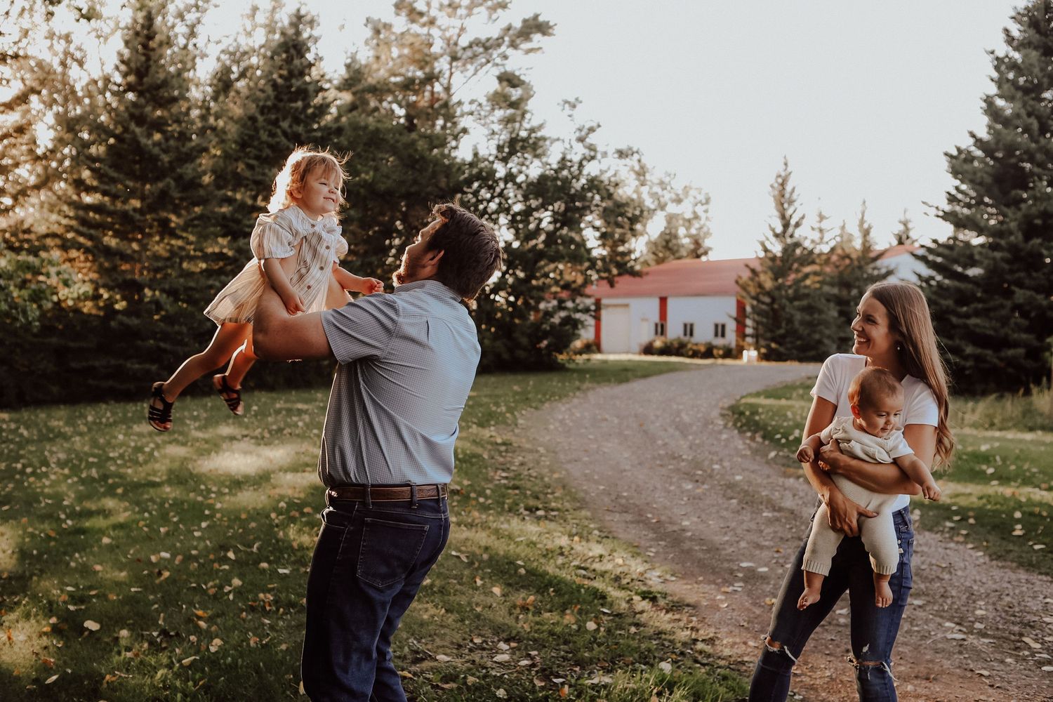 A joyful family plays outdoors at golden hour, father lifting toddler while mother holds baby near a red barn.