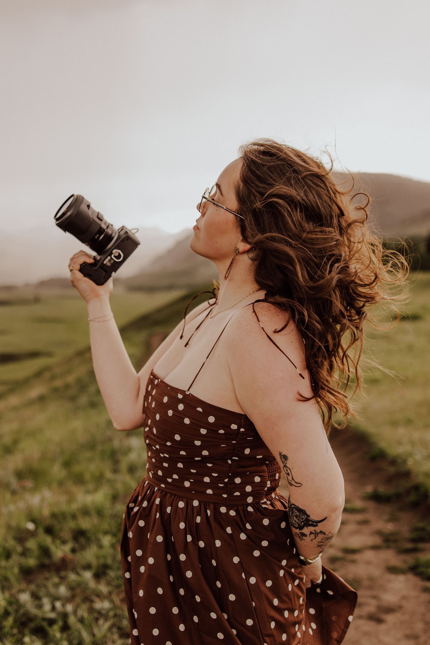 Photographer in brown polka dot dress takes photos against rolling hills at sunset.
