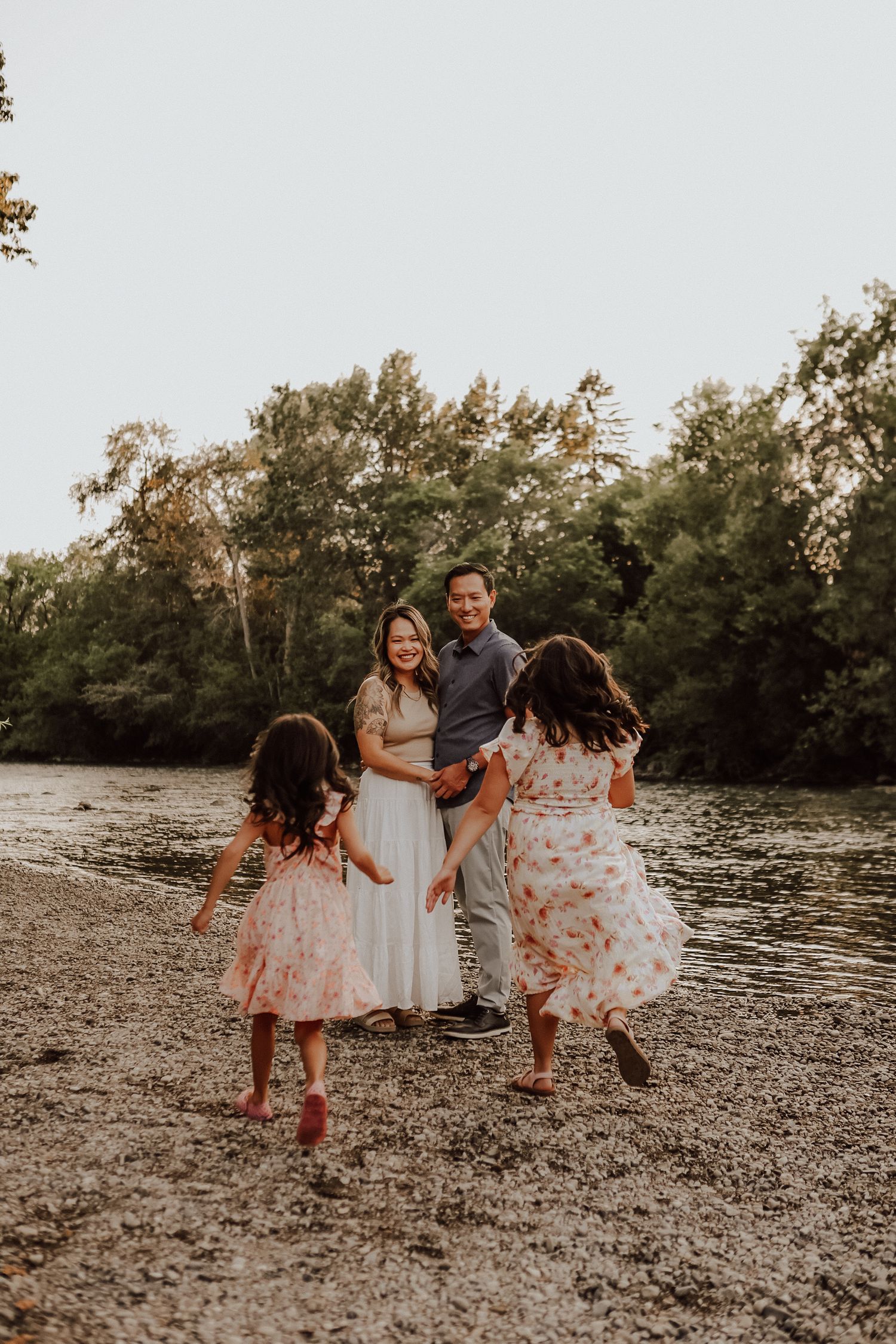 Family of four enjoying a golden hour photoshoot by a riverside, with two girls in floral dresses running toward their parents.