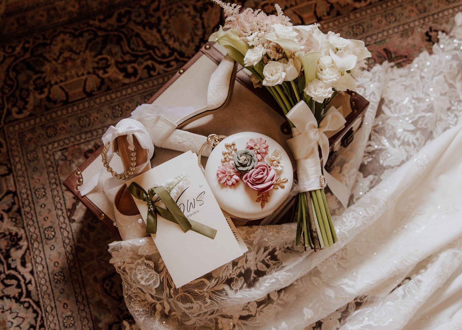 Elegant wedding flat lay with floral cake, white bouquet, bridal shoes, and vows card on ornate rug.