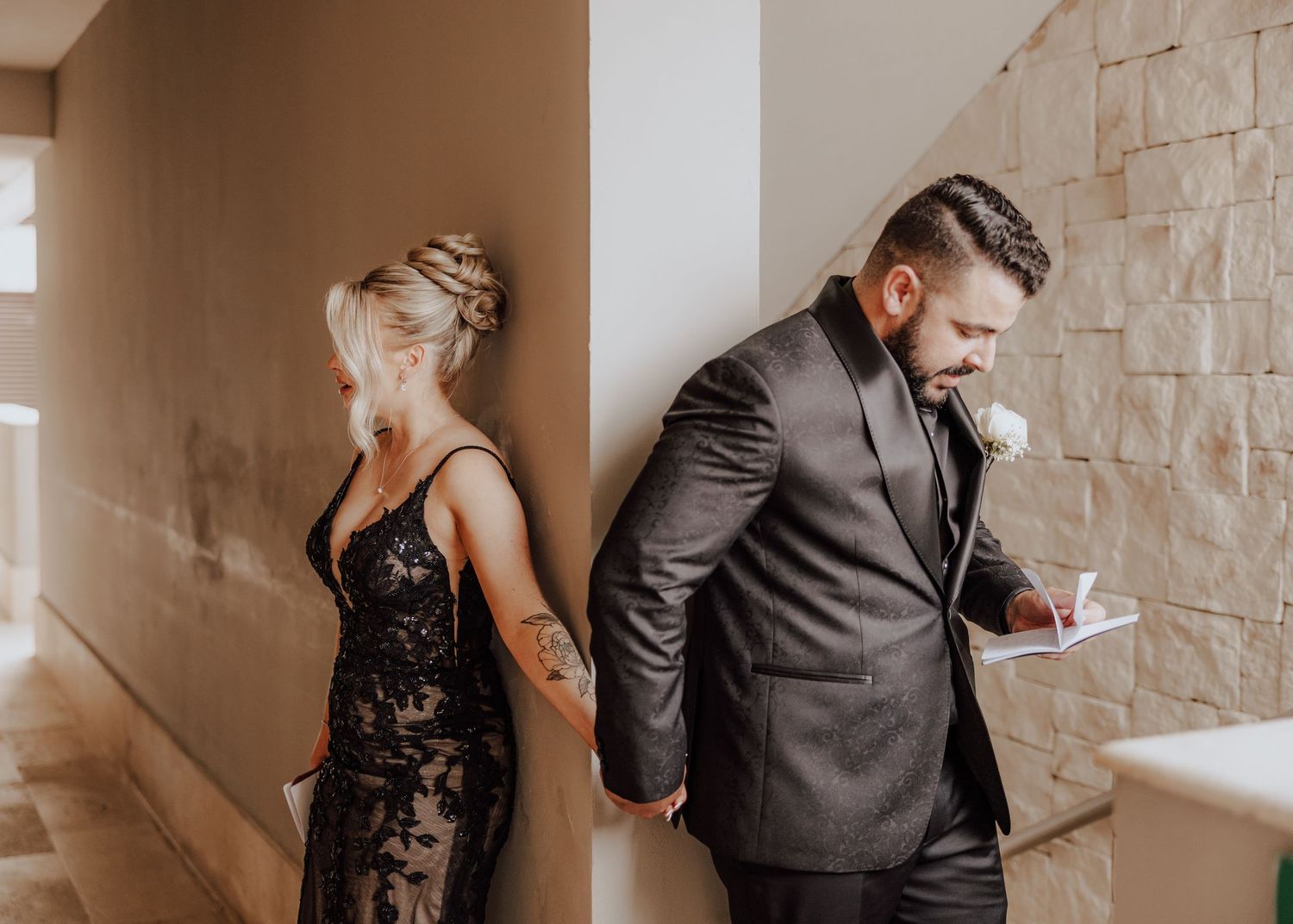A romantic couple in formal attire shares intimate moments against a stone wall in a wedding venue.