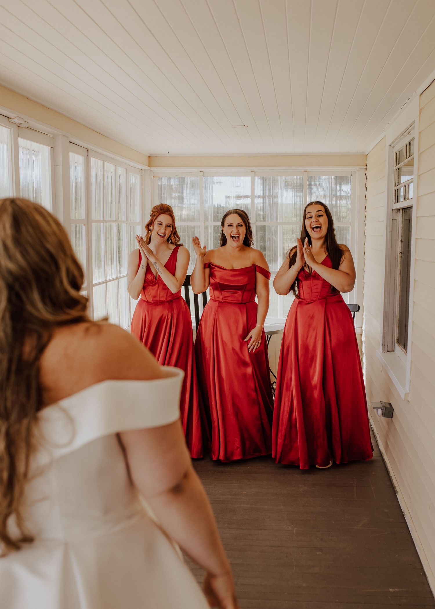 A wedding party in red bridesmaid dresses and white bridal gown laughing together in a bright hallway.