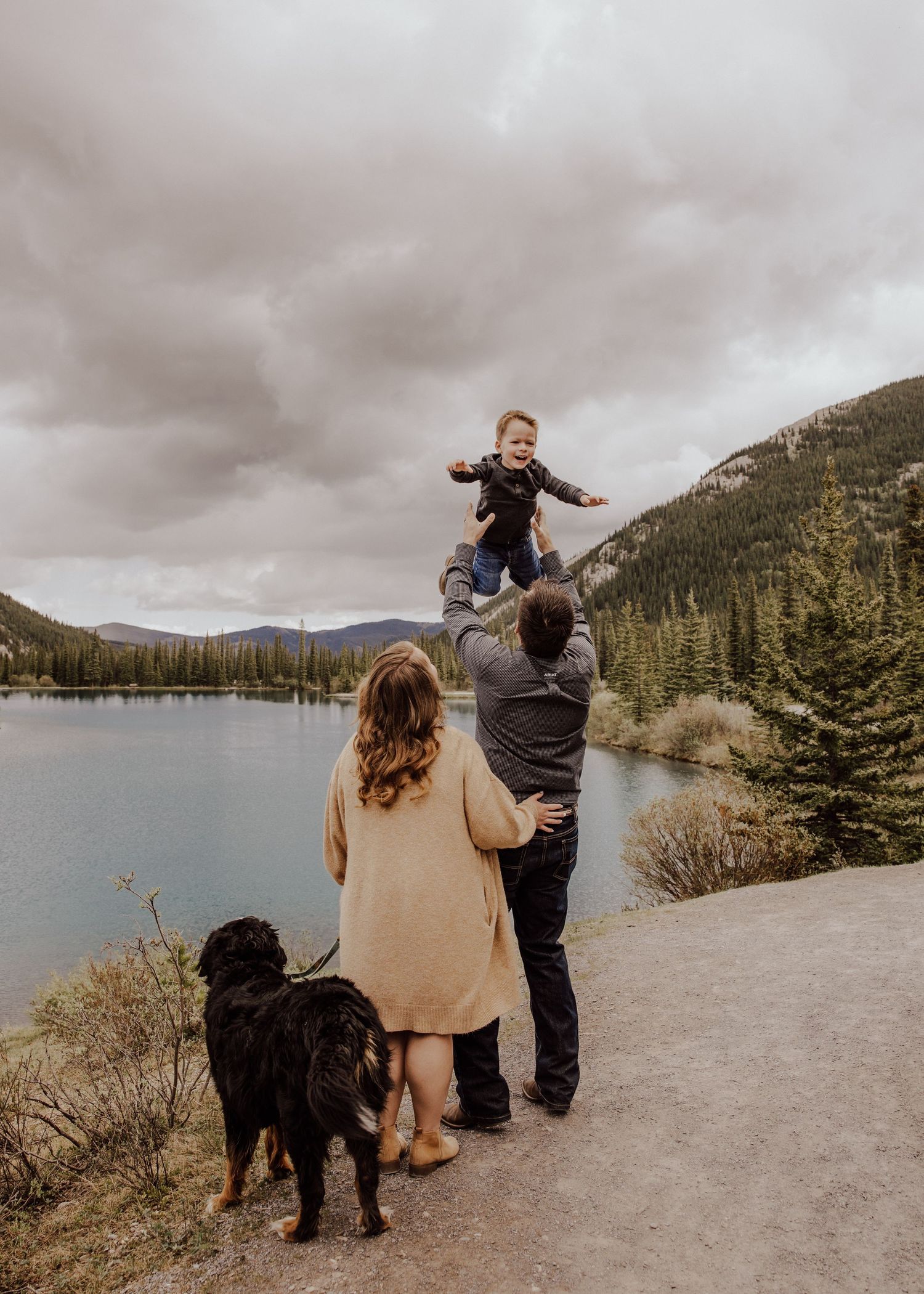Family with dog admiring mountain lake view, parent lifting toddler in air, scenic forest backdrop.