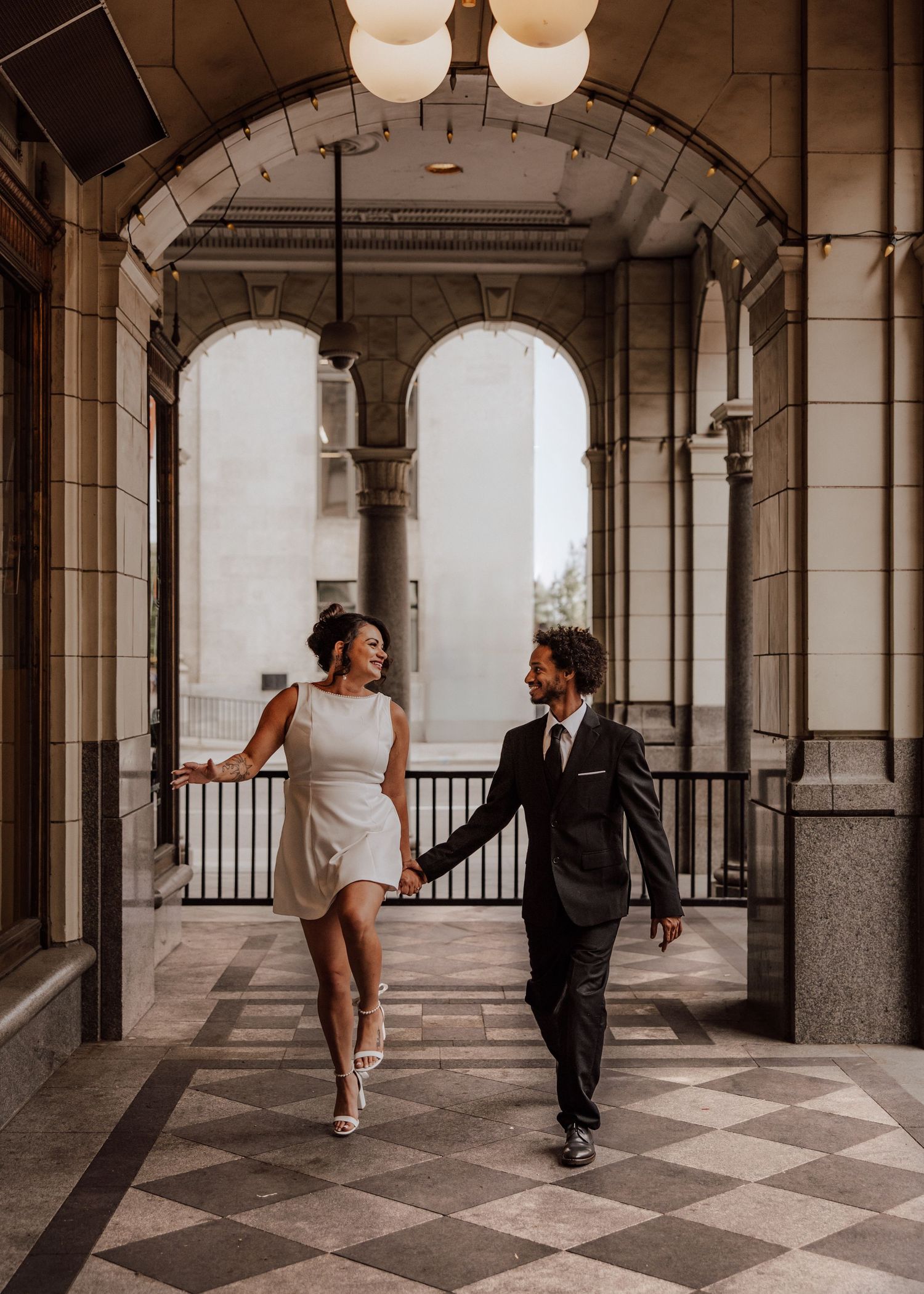 A couple dances playfully in an elegant columned hallway with a checkered floor pattern.