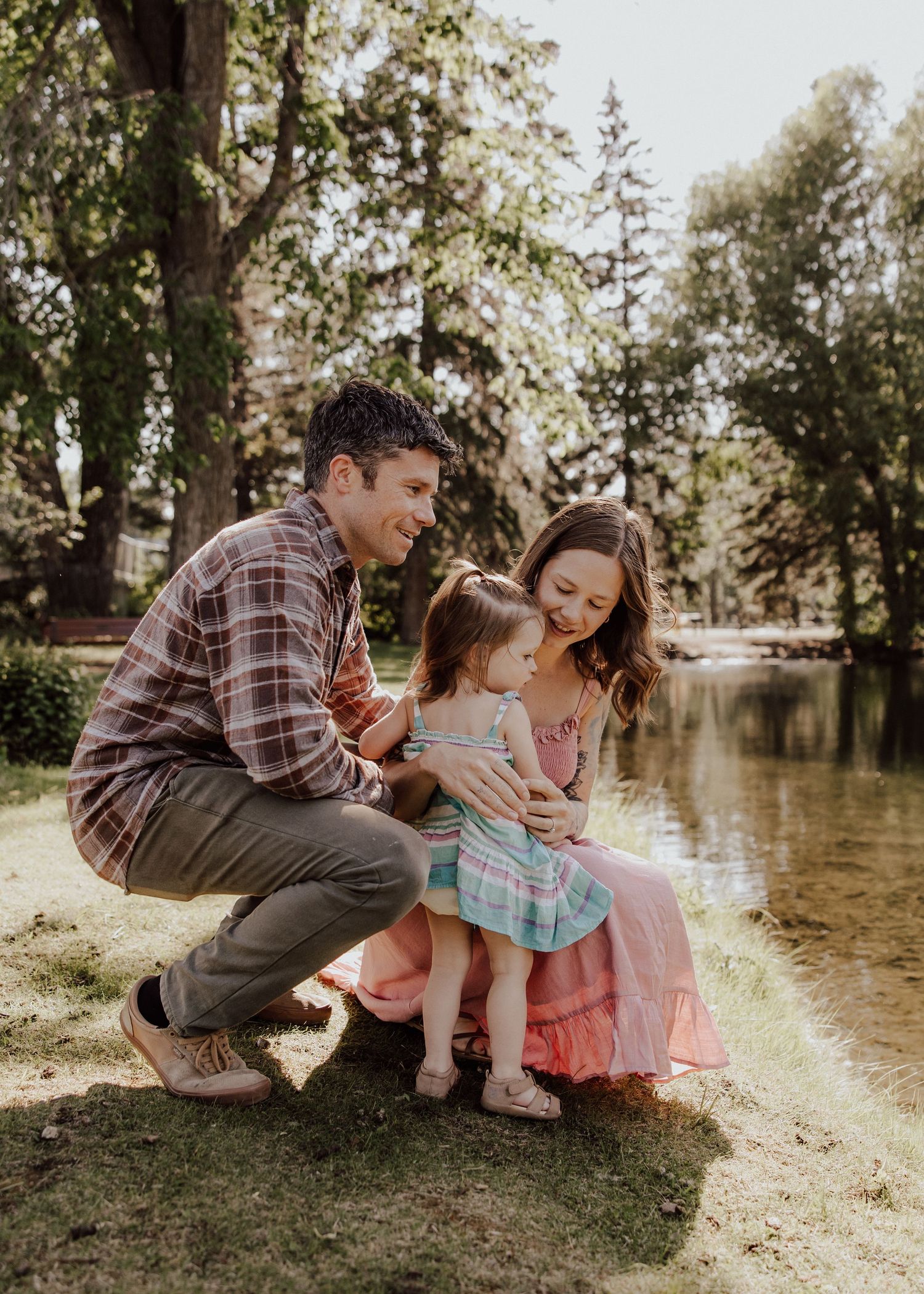 A family of three shares a tender moment by a sunlit pond, with parents smiling at their toddler daughter outdoors.