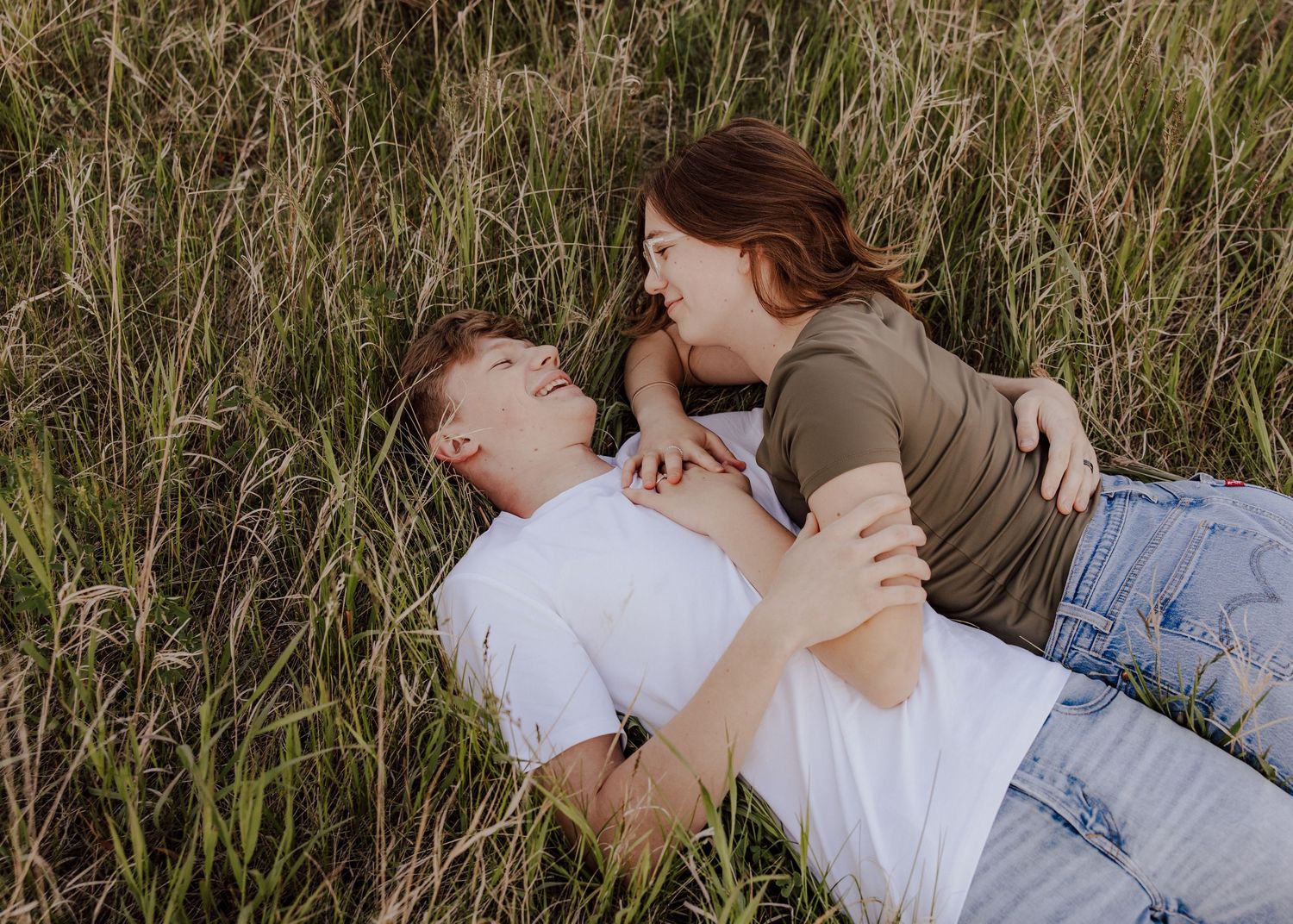 A couple lying in tall green grass, gazing at each other romantically during an outdoor couples photo session.