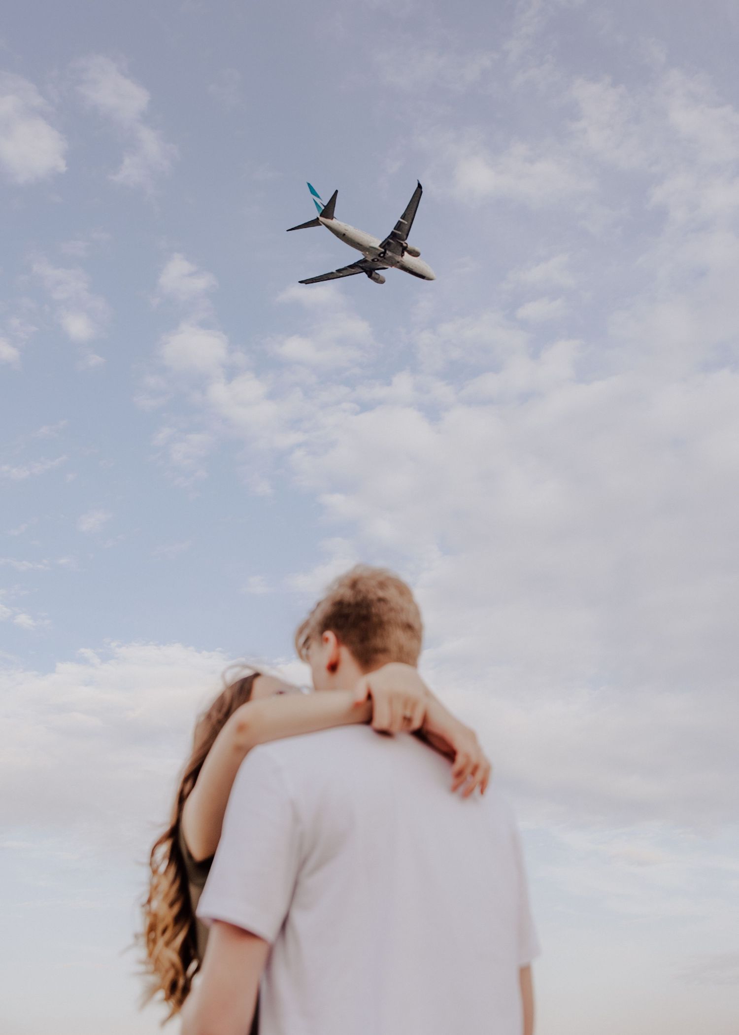Couple embracing outdoors, gazing up at a commercial airplane flying through a partly cloudy blue sky.