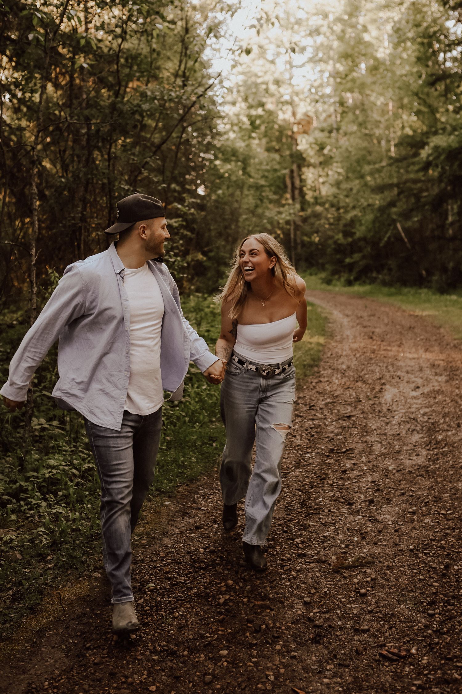 A couple playfully walks down a forest trail covered in fallen leaves.