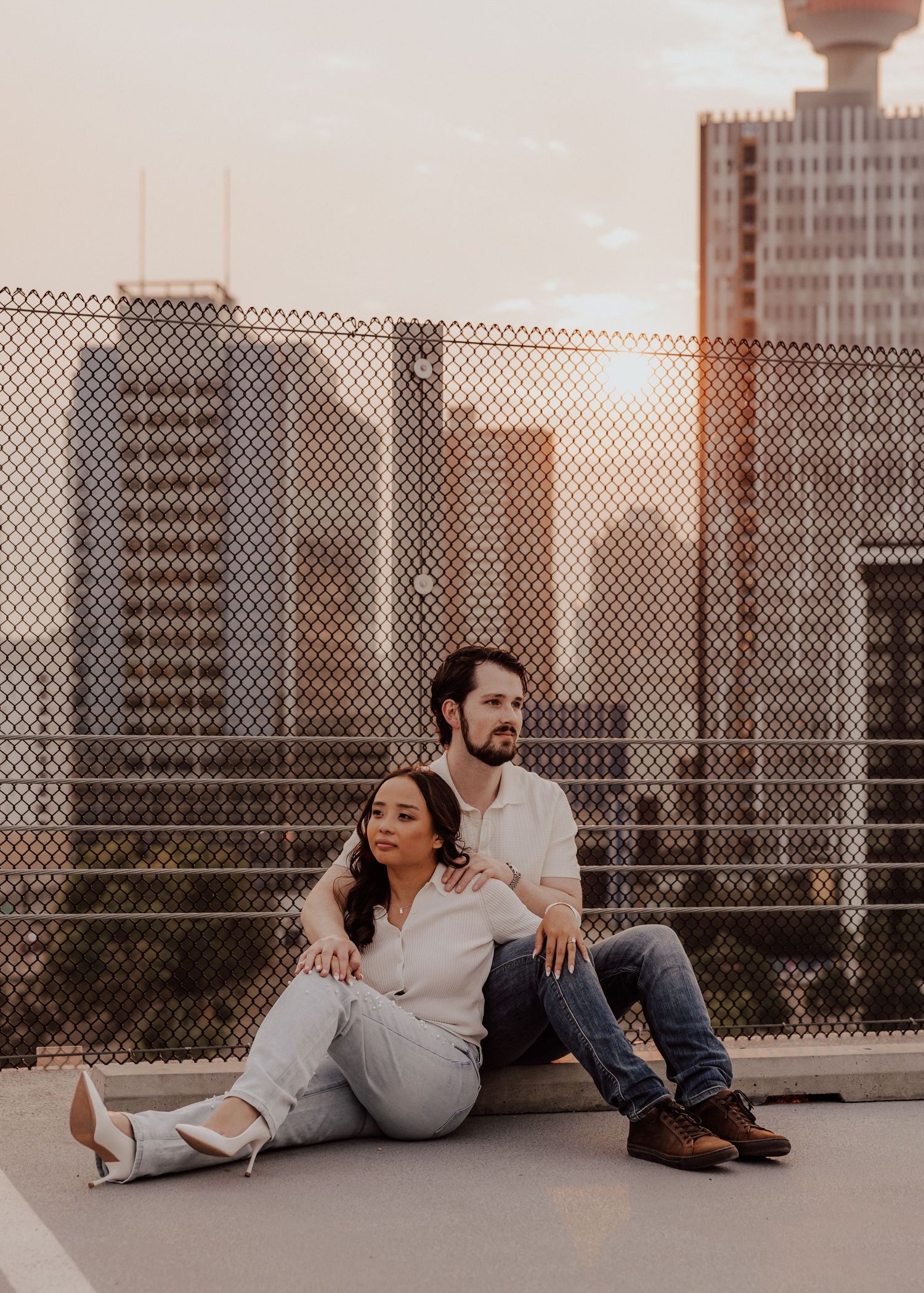 Couple in casual attire sitting against urban rooftop fence during sunset.