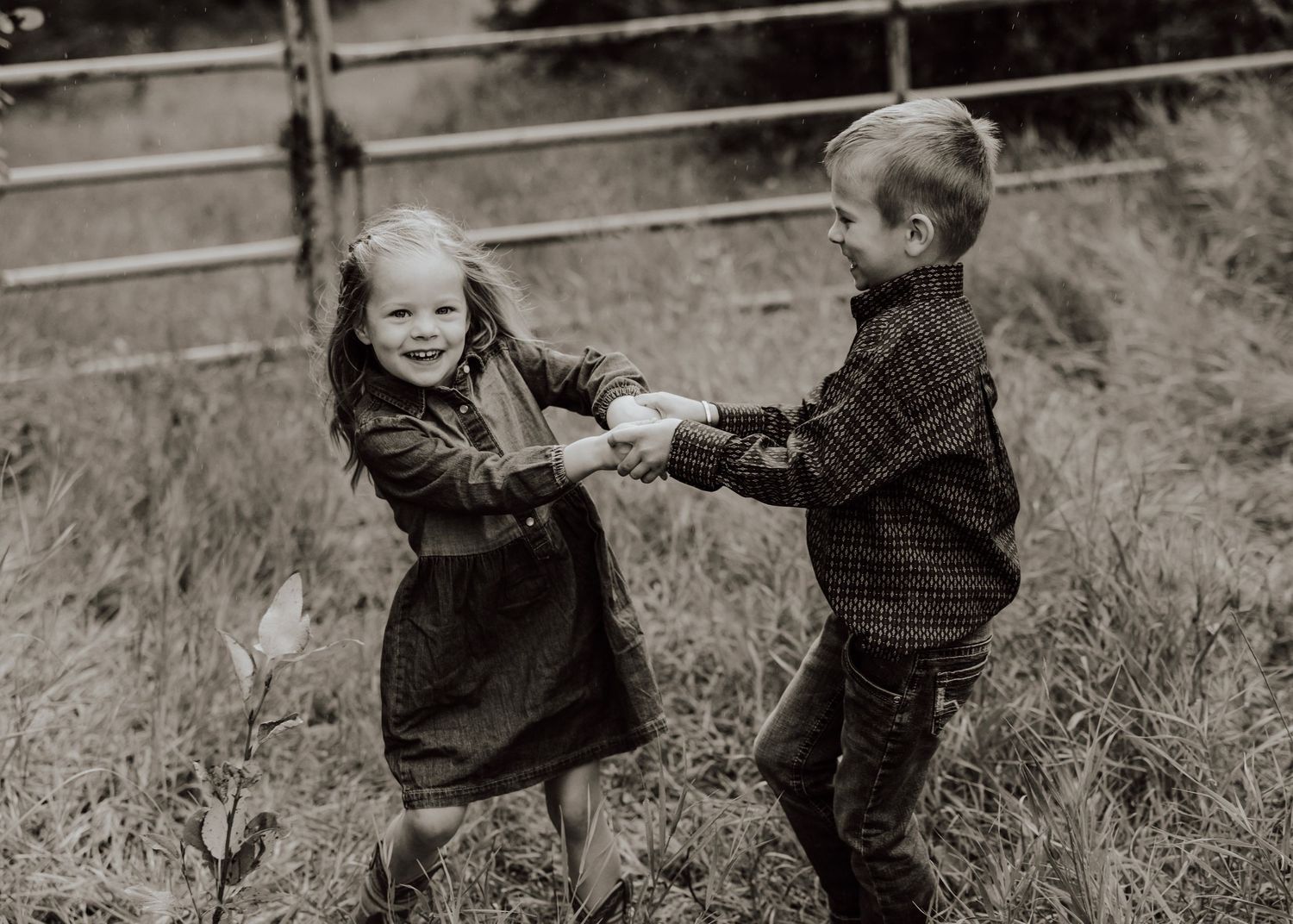 Two young children joyfully hold hands and spin together in a grassy field in a black and white photo.