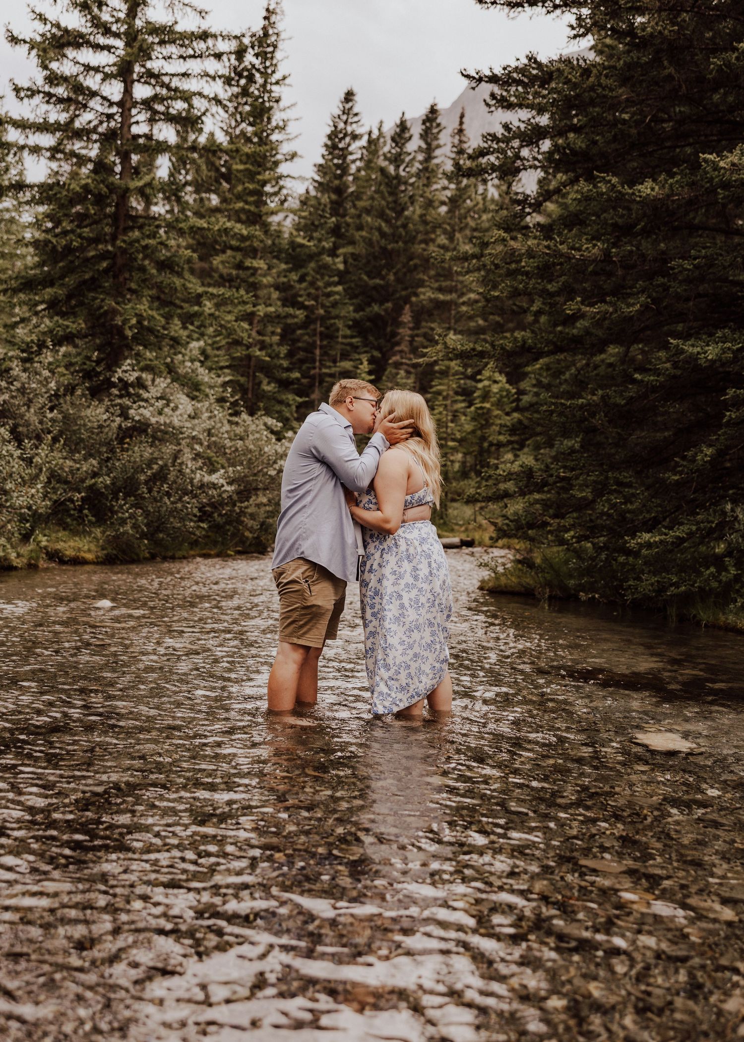 A couple shares an intimate moment while standing in shallow river water surrounded by pine trees.