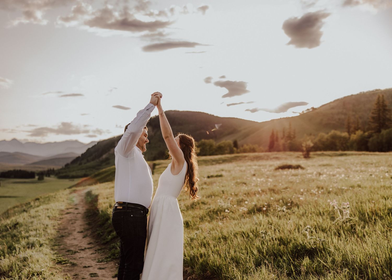 A couple in wedding attire walks through a scenic mountain meadow at sunset with dramatic clouds overhead.