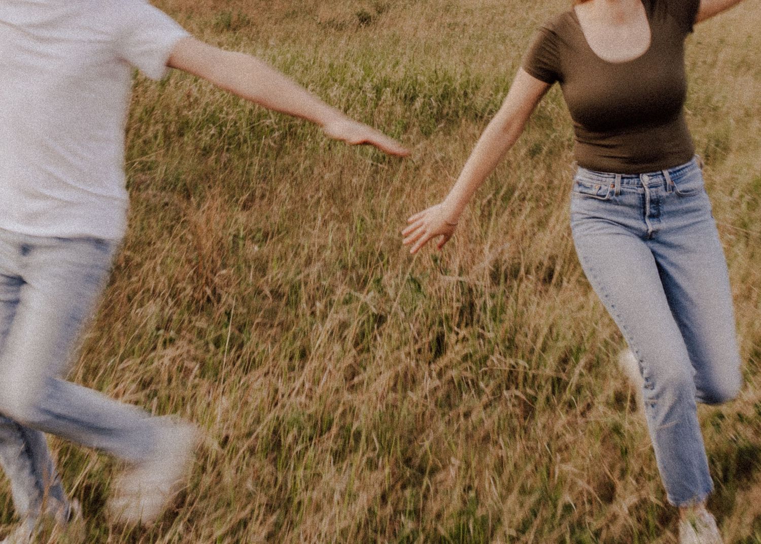 A couple runs through a golden grass field, reaching for each other's hands in a playful outdoor moment.
