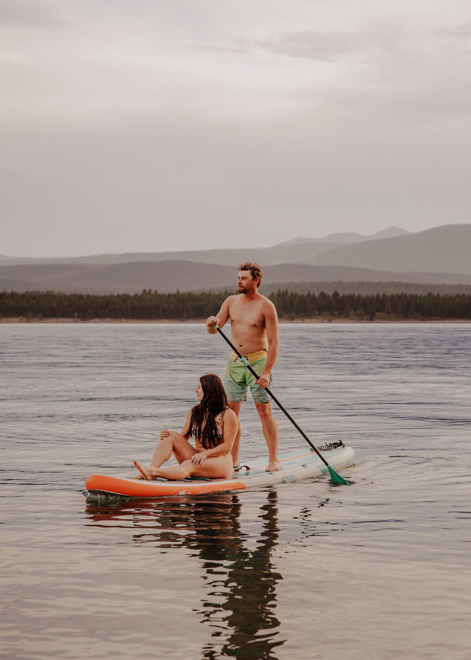 A couple enjoys paddleboarding on a calm lake with mountain views, man standing with paddle, woman sitting on board.