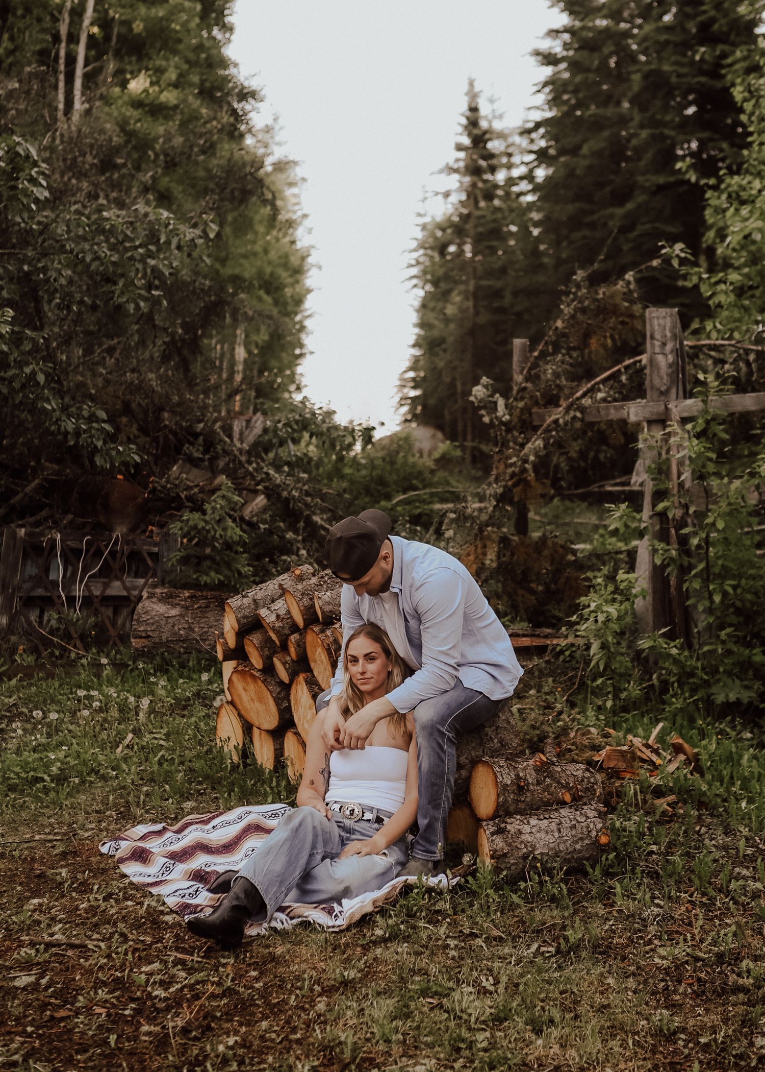 A couple relaxes on a blanket near stacked firewood in a forest setting.