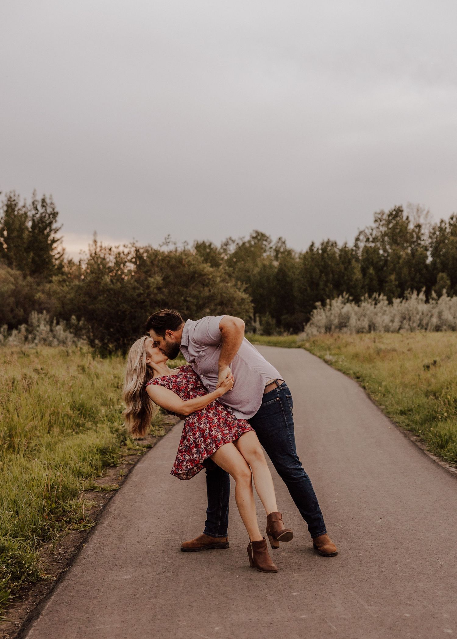 A couple in casual attire performs a romantic dip pose sequence on a rural pathway at sunset.