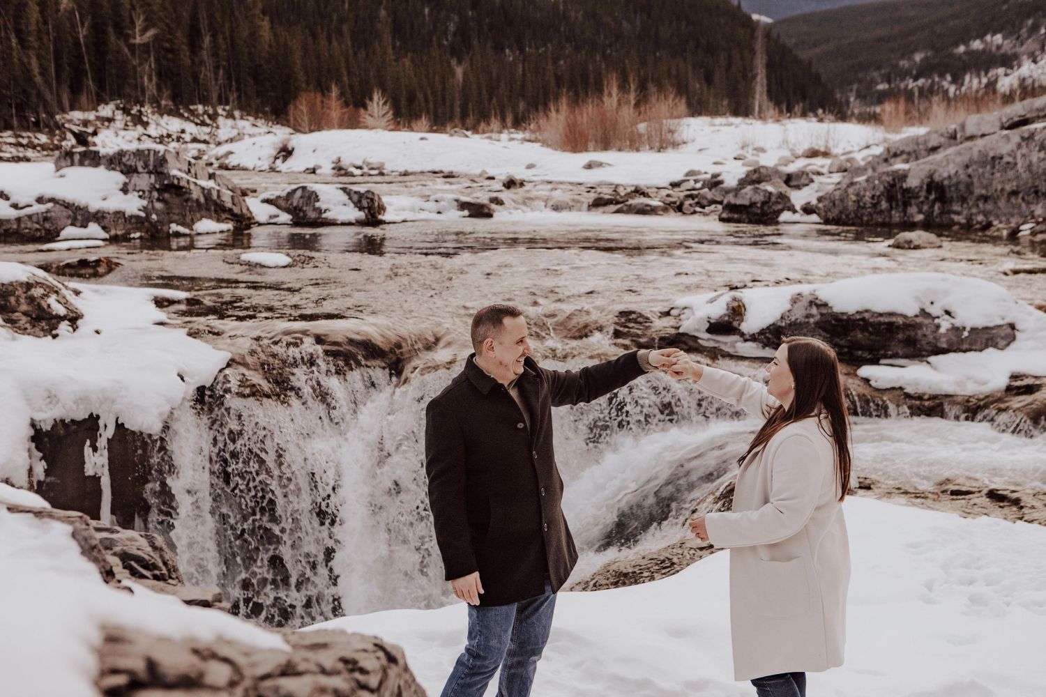 A couple walks together in a snowy winter landscape with mountains and trees in the background.