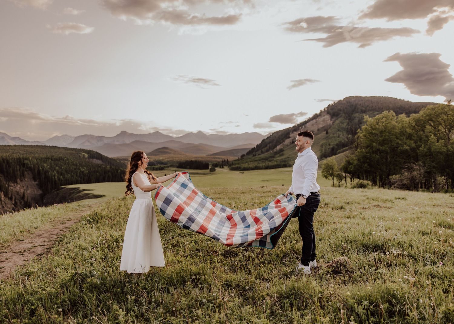 Couple holding a colorful plaid blanket in a green meadow with mountain backdrop at sunset.