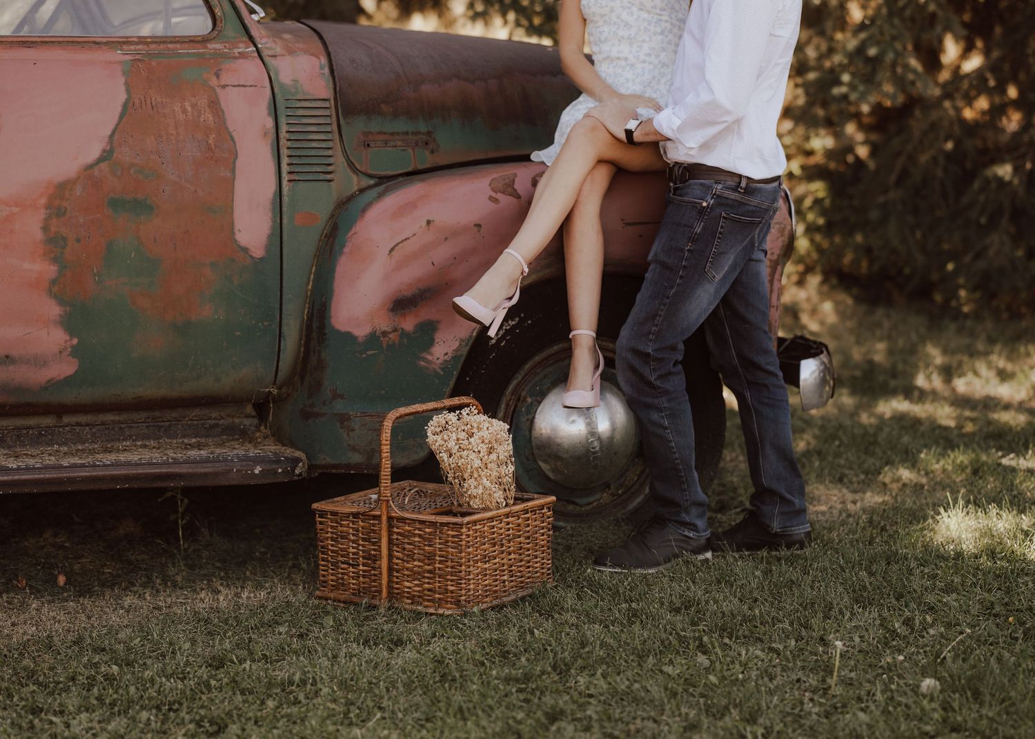 Couple posing near a rustic vintage car with a wicker picnic basket on green grass in a romantic outdoor setting.