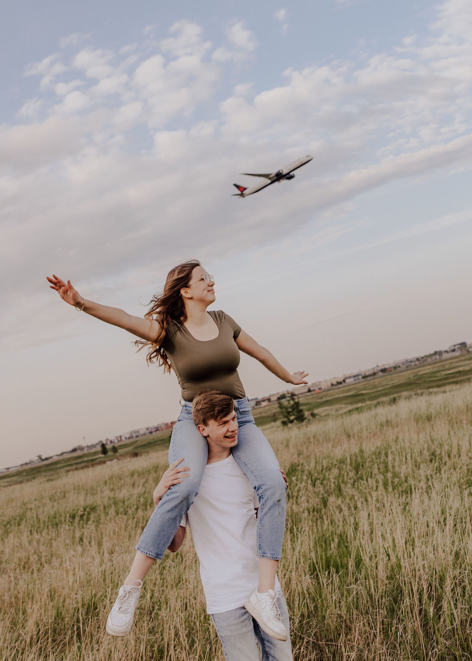 A young couple joyfully poses in a field, woman on man's shoulders with arms spread wide, airplane flying overhead.