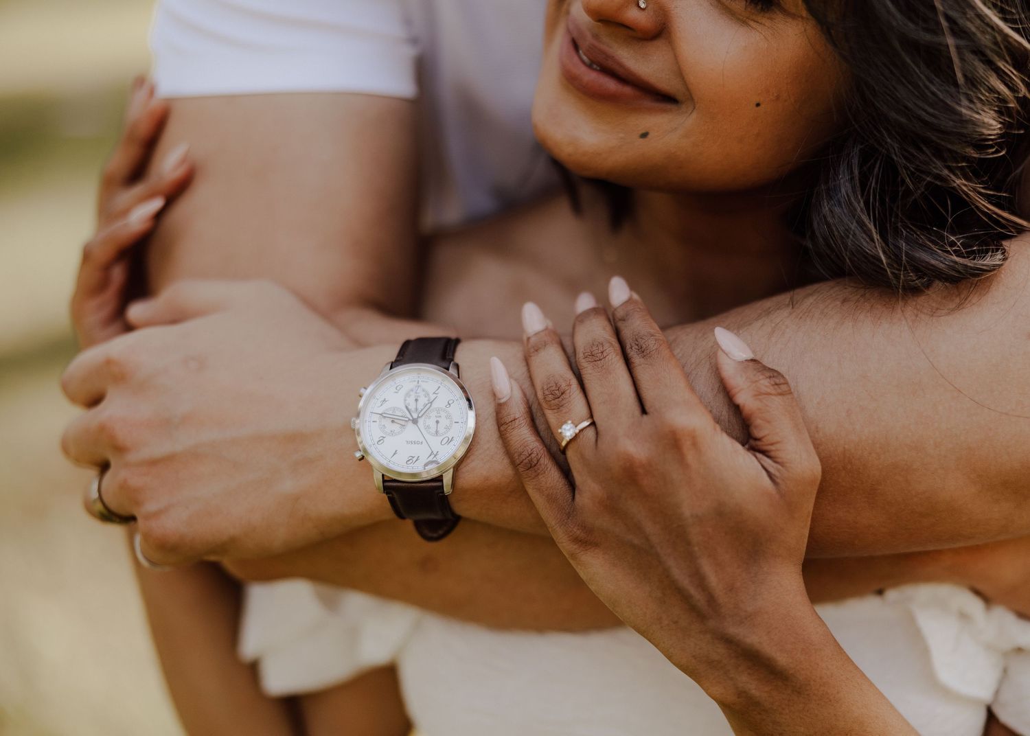 Close-up view of hands wearing an elegant watch and engagement ring with a white dress in soft lighting.